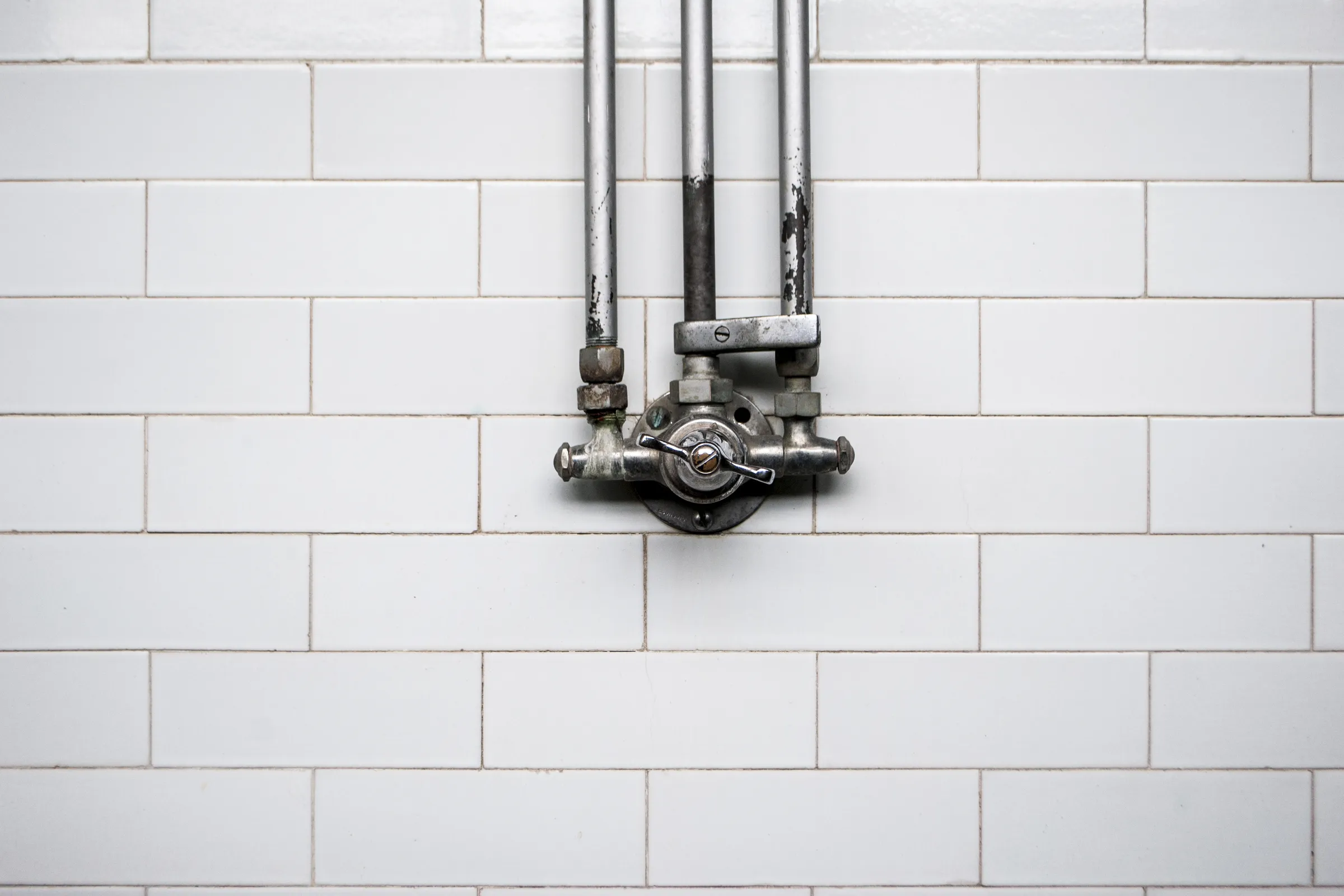A white tiled wall with the shower pipes and mixer running on its surface.